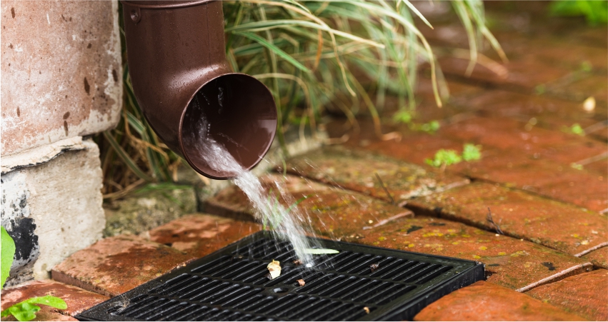 Rainwater flowing from a downspout onto a grate on a brick surface.