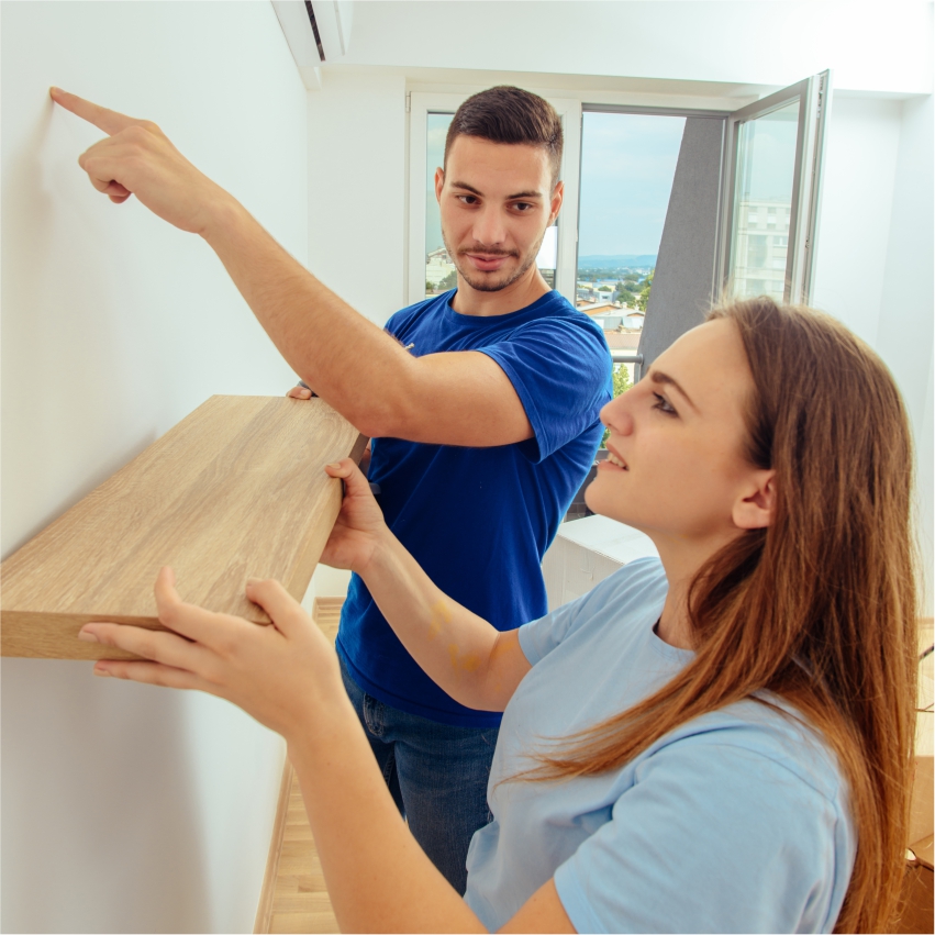 A man and woman pointing at a wooden shelf in a room.