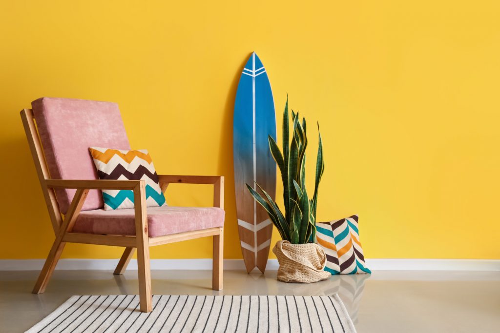 A pink chair with a surfboard against a yellow wall.