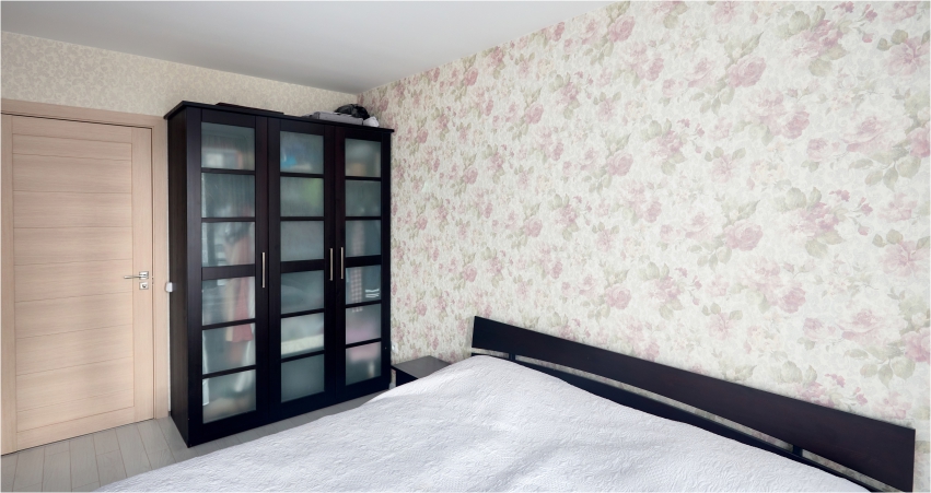 A modern bedroom with a large bed, floral wallpaper, and a glass door wardrobe.