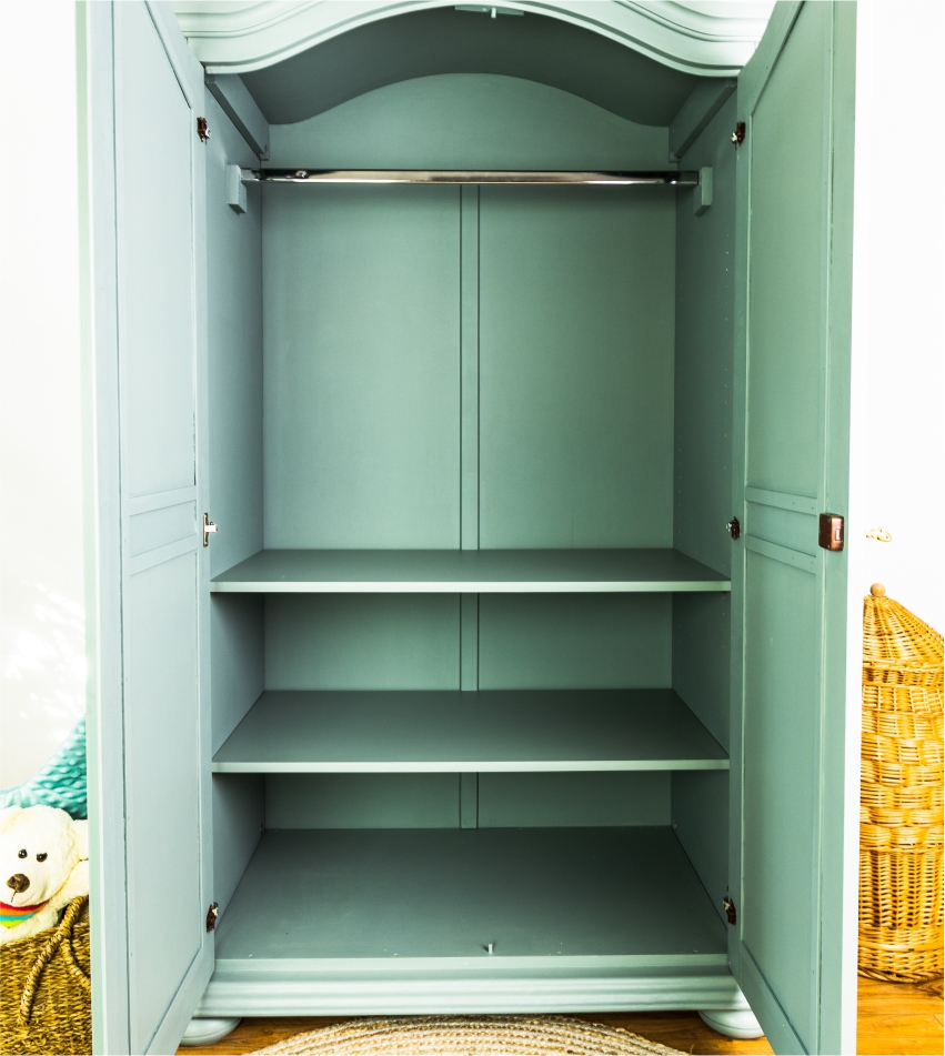 An open empty wardrobe with shelves against a white wall, with a portion of a wicker basket and a soft toy visible to the left.