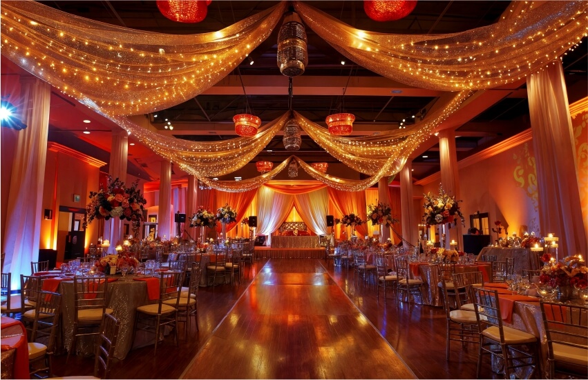 Wedding venue with a decorated mandap, draped ceiling lights, and a glossy dance floor. 