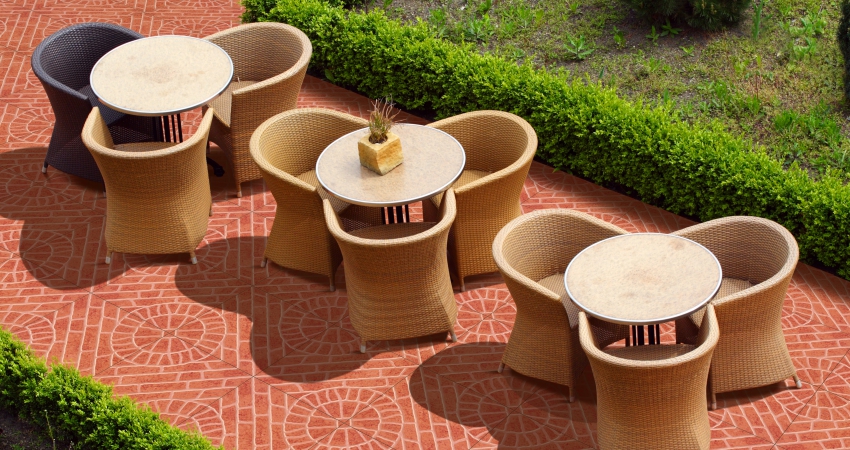 Outdoor patio seating area with round tables, wicker chairs, and hedges on red tile flooring.